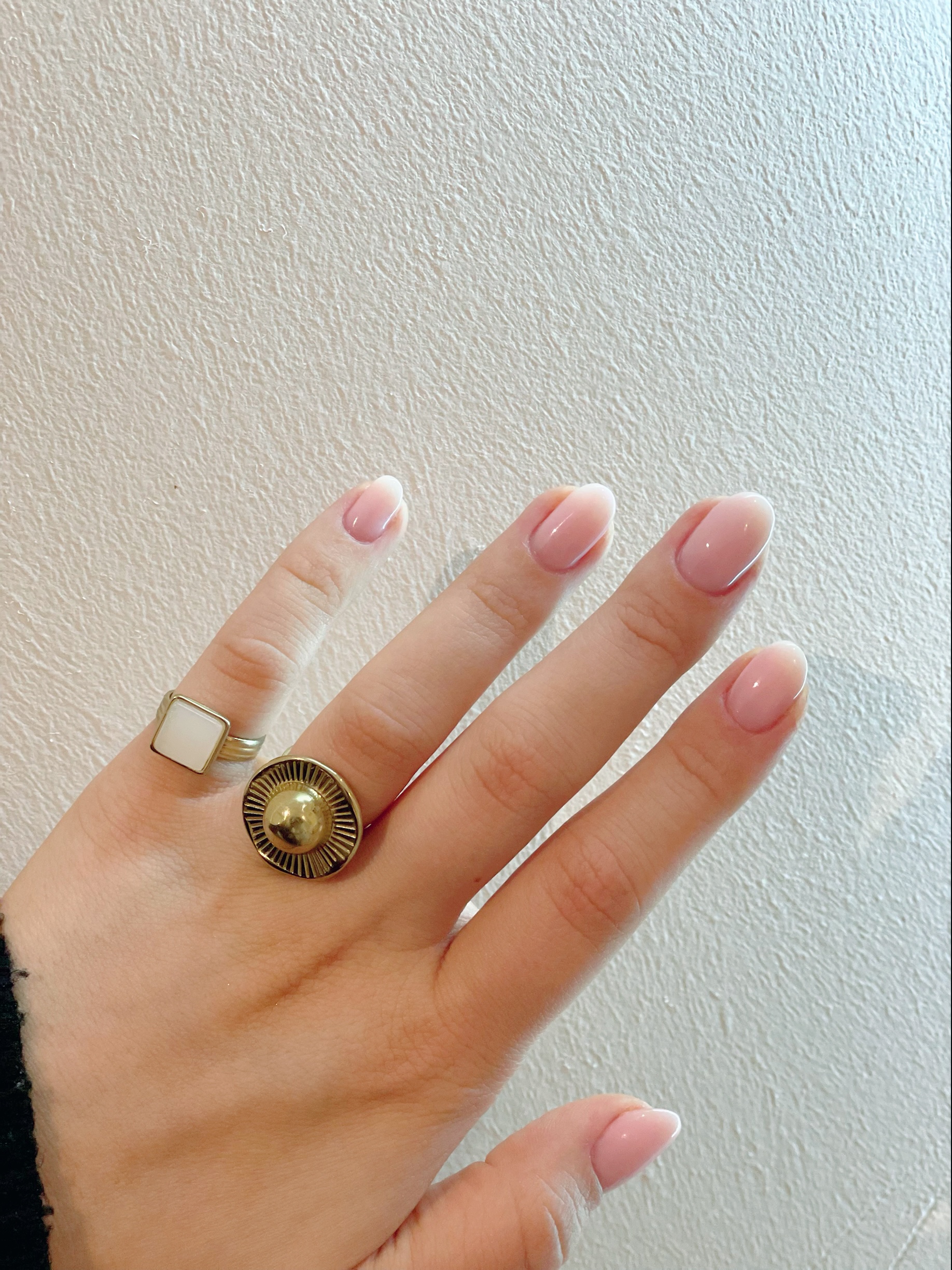 Zachtroze French nagels, een hand met gouden ringen tegen een witte muur